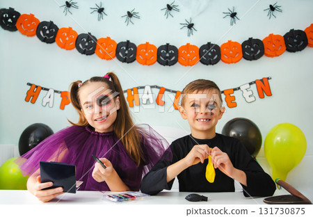 Two kids getting ready for Halloween. One girl applies makeup while boy inflates balloon. Two kids getting ready for Halloween. One girl applies makeup while boy inflates balloon. 131730875