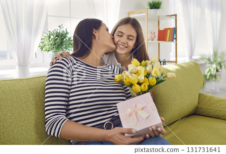 Child girl congratulating mother with flowers tulips and present box sitting on sofa at home. Child girl congratulating mother with flowers tulips and present box sitting on sofa at home. 131731641