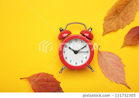 Red alarm clock, acorns and autumn leaves on yellow background. Daylight saving time end Red alarm clock, acorns and autumn leaves on yellow background. Daylight saving time end 131732305