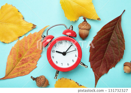 Red alarm clock, acorns and autumn leaves on blue background. Daylight saving time end Red alarm clock, acorns and autumn leaves on blue background. Daylight saving time end 131732327