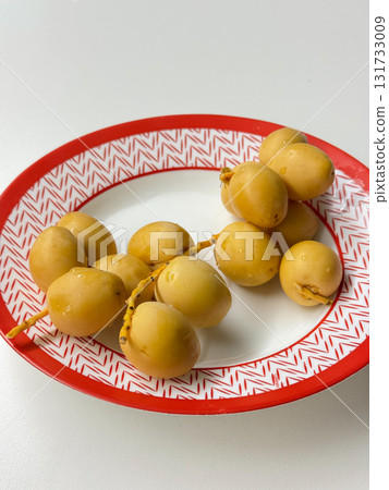 Fresh yellow dates with water drops lie on a white plate with a red geometric rim, illuminated by soft natural light 131733009