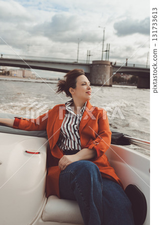 Brunette happy woman in casual sits in boat sailing along river in European city on sunny day Brunette happy woman in casual sits in boat sailing along river in European city on sunny day 131733613