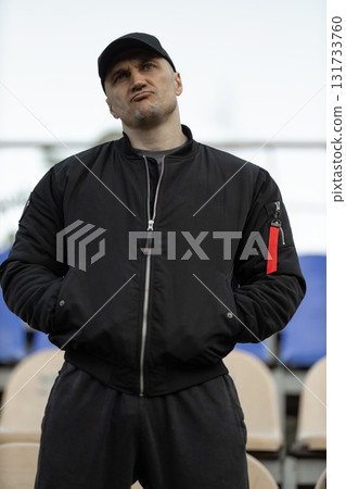 In an empty stadium man wearing black bomber jacket and cap stands with arms crossed expressing emotion while watching his team compete during game. In an empty stadium man wearing black bomber jacket and cap stands with arms crossed expressing emotion while watching his team compete during game. 131733760