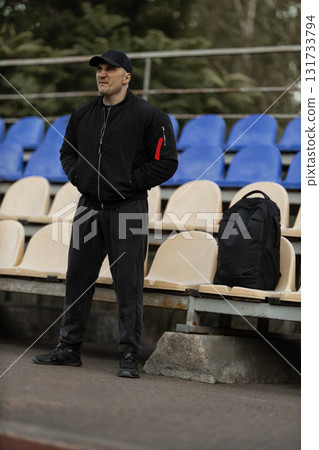 man stands in the empty stands wearing a black bomber jacket and cap focused on the unfolding sports game. His body language shows deep engagement with the match taking place. 131733794