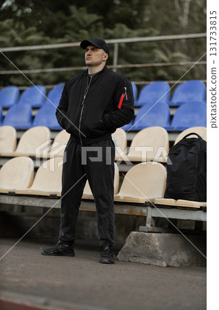 In an empty stadium a man dressed in a black bomber jacket and cap stands alone in the stands while watching an exciting game unfold. 131733815