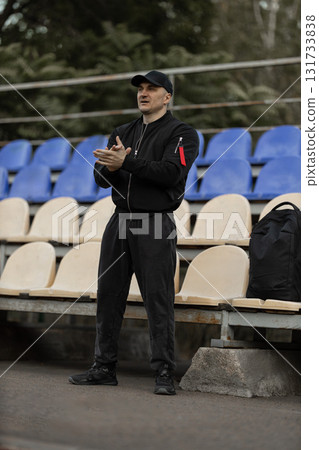 man wearing a black bomber jacket and cap stands in the empty stadium clapping and showing enthusiasm for the team during a game. The atmosphere is tense with anticipation. 131733838
