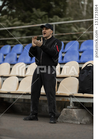 man stands in the empty stadium wearing a black bomber jacket and cap. He claps and reacts enthusiastically to the team's game showing his support for the players. man stands in the empty stadium wearing a black bomber jacket and cap. He claps and reacts enthusiastically to the team's game showing his support for the players. 131733864
