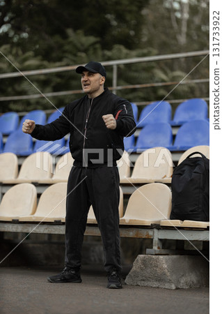 man wearing black bomber jacket and cap stands excitedly in empty stadium seats cheering on his team during intense game conveying strong emotions and enthusiasm. man wearing black bomber jacket and cap stands excitedly in empty stadium seats cheering on his team during intense game conveying strong emotions and enthusiasm. 131733922
