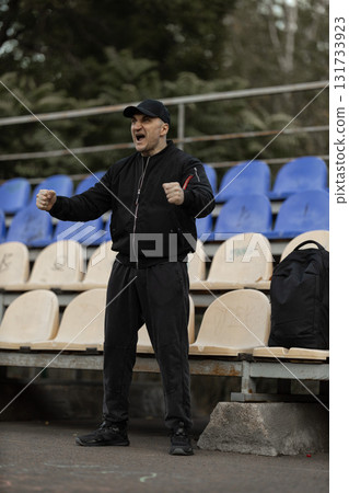 man wearing black bomber jacket and cap stands excitedly in empty stadium seats passionately reacting to team performance during match day clearly invested in action. man wearing black bomber jacket and cap stands excitedly in empty stadium seats passionately reacting to team performance during match day clearly invested in action. 131733923