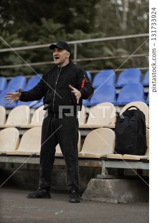 In a quiet stadium a man in a black bomber jacket and cap stands in the empty stands expressing strong emotions as he reacts to the game unfolding. His energy adds life to the scene. 131733924