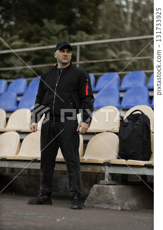 man wearing black bomber jacket and cap stands in empty stadium stands showing his reactions to ongoing team game with focused attention and emotion. 131733925