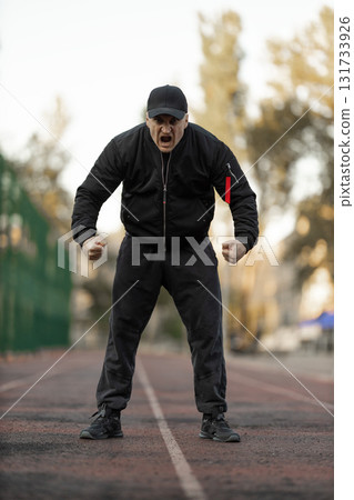 man in a black bomber jacket and athletic wear stands on a treadmill actively engaging in a workout at an outdoor gym during the day. His focused expression suggests dedication to fitness. 131733926