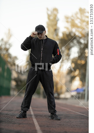 man dressed in a black bomber jacket and sportswear stands focused on a treadmill. The setting is an outdoor gym with trees in the background. It is late afternoon providing a warm light. man dressed in a black bomber jacket and sportswear stands focused on a treadmill. The setting is an outdoor gym with trees in the background. It is late afternoon providing a warm light. 131733930