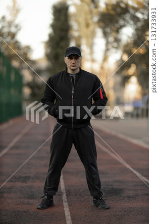 man in a black bomber jacket stands confidently on a treadmill while dressed in athletic clothes. He is at an outdoor gym where fitness enthusiasts often train during the early morning hours. 131733931