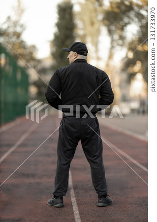 man dressed in a black bomber jacket and athletic clothing stands on a treadmill at an outdoor gym. The scene is set in the morning with soft sunlight illuminating the area. 131733970