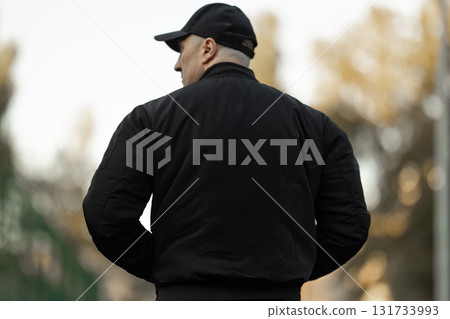 man wearing a black bomber jacket and sportswear stands on a treadmill in an outdoor setting. The environment features blurred trees in the background showing an active and healthy lifestyle. man wearing a black bomber jacket and sportswear stands on a treadmill in an outdoor setting. The environment features blurred trees in the background showing an active and healthy lifestyle. 131733993