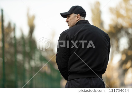 man wearing a black bomber jacket and sportswear stands on a treadmill. He appears focused and determined in an outdoor fitness space surrounded by greenery during the day. 131734024