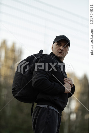 man wearing a black bomber jacket and cap stands with a backpack on his shoulder. He looks towards the distance with a serious expression in a natural outdoor location during the day. 131734051