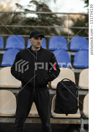 man wearing a black bomber jacket and cap stands in an empty stadium tribune. He has a backpack next to him and looks out over the seats in daylight. 131734130