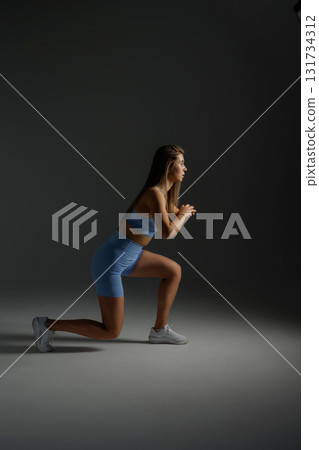 Fitness model performing a lunge exercise in a dimly lit studio showcasing strength and determination 131734312