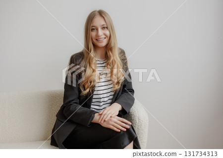 Young woman in a professional outfit poses confidently on a light sofa in a modern office setting during the day Young woman in a professional outfit poses confidently on a light sofa in a modern office setting during the day 131734313