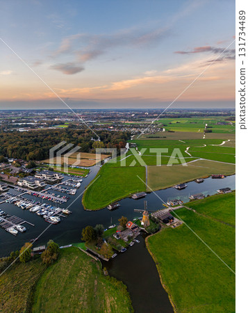 Aerial view of rural marina boats docked in winding waterways, windmill near canal, scattered buildings, and village framed by green fields and warm sunset sky in a tranquil Dutch style layout. Aerial view of rural marina boats docked in winding waterways, windmill near canal, scattered buildings, and village framed by green fields and warm sunset sky in a tranquil Dutch style layout. 131734489