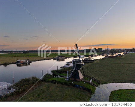 Aerial view of Dutch countryside dark green windmill near calm canal, houseboats and small buildings line banks, flat fields stretch under clear gradient sky with distant windmill at sunset. Aerial view of Dutch countryside dark green windmill near calm canal, houseboats and small buildings line banks, flat fields stretch under clear gradient sky with distant windmill at sunset. 131734490