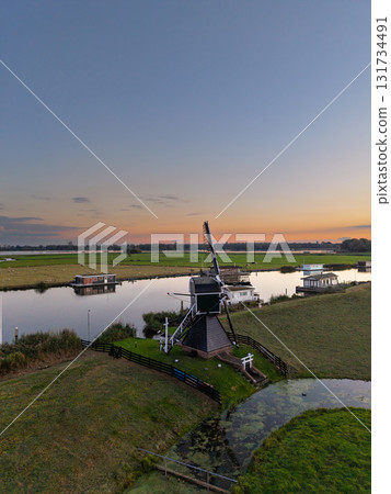 Aerial view of Dutch countryside dark green windmill with white accents near calm canal, houseboats and fences line grassy banks, sunset sky reflects warm hues across flat, tranquil landscape. Aerial view of Dutch countryside dark green windmill with white accents near calm canal, houseboats and fences line grassy banks, sunset sky reflects warm hues across flat, tranquil landscape. 131734491