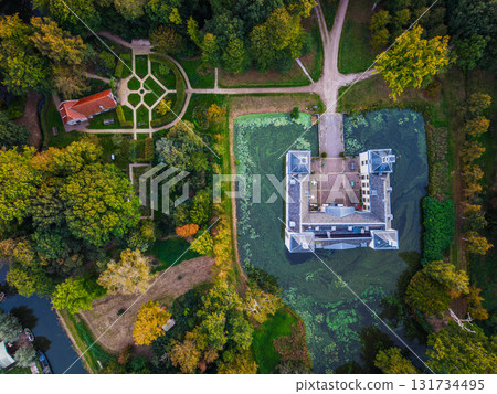 Aerial view of historic estate moated rectangular mansion with gabled roofs and courtyard, formal garden with radial paths and red roofed pavilion, all framed by dense forest and dirt trails. Aerial view of historic estate moated rectangular mansion with gabled roofs and courtyard, formal garden with radial paths and red roofed pavilion, all framed by dense forest and dirt trails. 131734495
