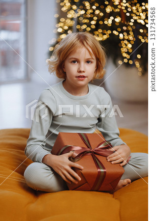 Child with gift box sitting indoors near Christmas tree lights, front view 131734568