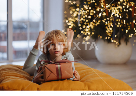 Smiling girl in pajamas holding gift box lying on cushion near Christmas tree with lights Smiling girl in pajamas holding gift box lying on cushion near Christmas tree with lights 131734569
