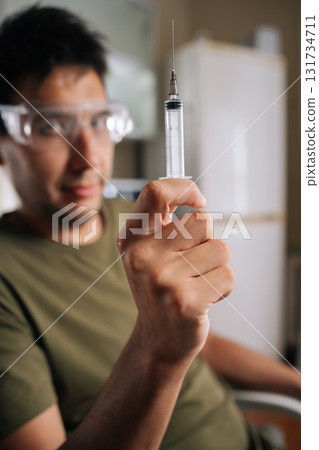 Selective focus portrait of chemist wearing safety glasses carefully preparing fertilizer using syringe, ensuring precise measurements for optimal plant growth and healthy harvest, looking at camera. 131734711