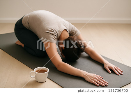 AI generated peaceful image of a person practicing child pose on yoga mat with a cup of tea next to them. Soothing, minimalist setting perfect for wellness, relaxation, and mindfulness content. 131736700