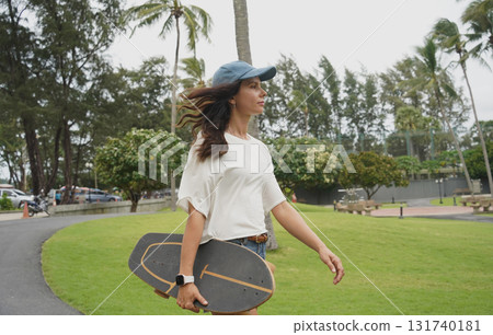 A woman is walking at the city park while carrying a skateboard in her hands 131740181