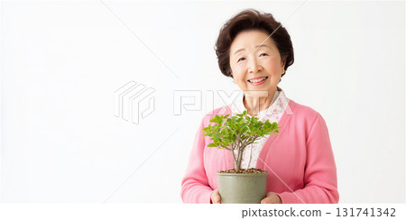 一位身穿粉紅色開襟衫的老年婦女,手裡拿著一盆盆栽,笑容燦爛,露出牙齒。 一位身穿粉紅色開襟衫的老年婦女,手裡拿著一盆盆栽,笑容燦爛,露出牙齒。 131741342