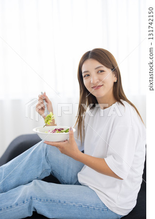 Young woman sitting on sofa and eating vegetable salad 131742150
