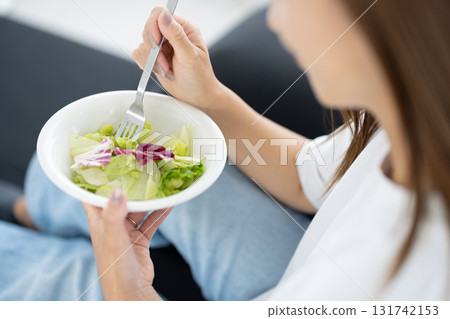 Young woman sitting on sofa and eating vegetable salad 131742153