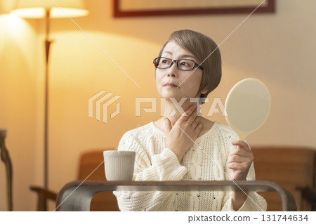 Senior woman looking at a hand mirror in the living room at night Senior woman looking at a hand mirror in the living room at night 131744634