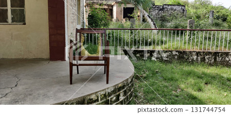 A wooden chair with a damaged seat sitting on a porch near a railing and overgrown yard 131744754