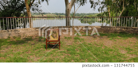 A chair on the grass near a railing overlooking a lake surrounded by trees and greenery 131744755