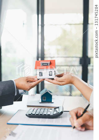 Close up of real estate agent handing house model to client with calculator and contract paperwork on desk, concept of mortgage loan, home ownership, property investment and housing agreement. Close up of real estate agent handing house model to client with calculator and contract paperwork on desk, concept of mortgage loan, home ownership, property investment and housing agreement. 131745284