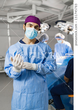 A surgeon in a surgical mask and scrubs prepares for surgery in a modern operating room with other medical staff. A surgeon in a surgical mask and scrubs prepares for surgery in a modern operating room with other medical staff. 131745783