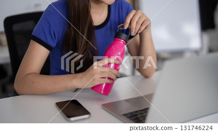 Asian adult woman relaxing at modern office desk staying hydrated with water bottle casual attire enjoying healthy break remote work setup 131746224