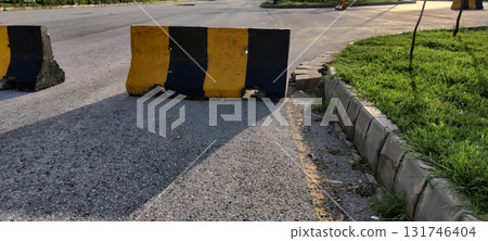 Road safety barrier painted in black and yellow stripes on asphalt with curb and grass in gentle afternoon lighting 131746404