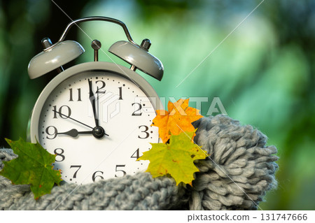 Classic alarm clock with autumn leaves nestled in a cozy gray scarf, time concept. 131747666