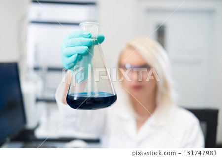 Blonde woman in rubber gloves holding a flask with blue solution with HPLC equipment in a molecular biology laboratory Blonde woman in rubber gloves holding a flask with blue solution with HPLC equipment in a molecular biology laboratory 131747981