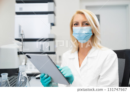 Blonde woman wearing a protective lab mask and gloves writing results of chemical experiments on a tablet in a fully equipped laboratory with flasks, beakers, and HPLC equipment. Blonde woman wearing a protective lab mask and gloves writing results of chemical experiments on a tablet in a fully equipped laboratory with flasks, beakers, and HPLC equipment. 131747987