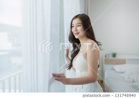 Young woman standing by window, enjoying bright morning sunlight, with sheer curtains in cozy bedroom for peaceful atmosphere and intimate moment Young woman standing by window, enjoying bright morning sunlight, with sheer curtains in cozy bedroom for peaceful atmosphere and intimate moment 131748940