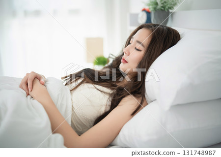 Young woman resting peacefully in a bright bedroom with soft natural light, feeling serene and relaxed while lying on a comfortable bed 131748987