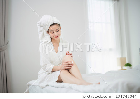 Relaxed woman in white robe applying lotion to skin after bath in bright bedroom with natural light and soft bedding for wellness and self-care concept Relaxed woman in white robe applying lotion to skin after bath in bright bedroom with natural light and soft bedding for wellness and self-care concept 131749044